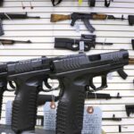 Pistols lined up on a display case and rifles on the wall of a gun store.